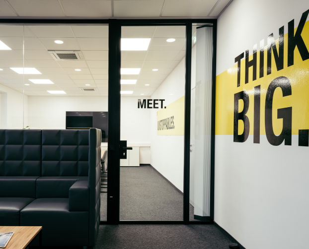 Besprechungsraum mit Glastür, schwarzem Loungesofa und motivierender Wandbeschriftung „THINK BIG.“