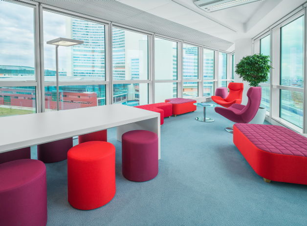 Lounge-Bereich mit roten und violetten Sitzmöbeln sowie weißen Tischen in einer hellen Ecke mit Rundumblick