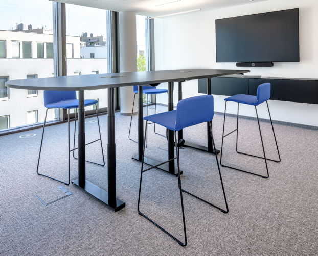 Besprechungstisch mit vier blauen Barhockern in einem lichtdurchfluteten Meetingraum mit Bildschirm