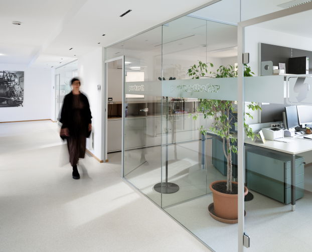 Langer, heller Büroflur mit Glaswänden, dahinter Arbeitsplätze und ein großer Bürotisch mit Monitoren und Pflanzen.