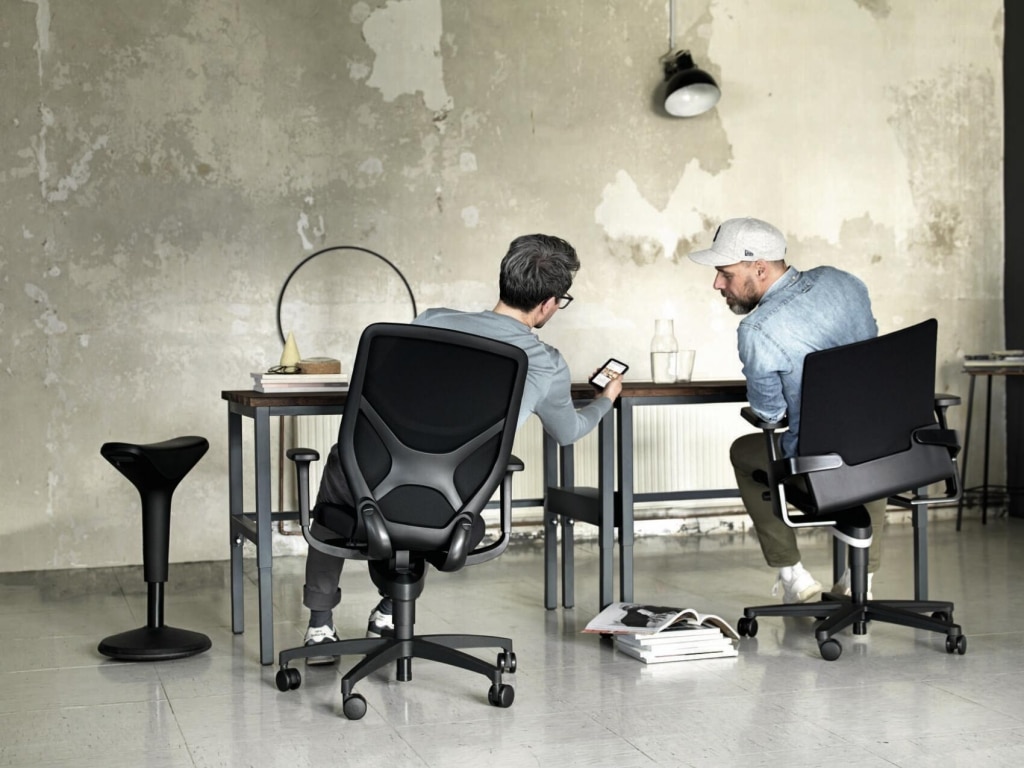 Open Office im Industriedesign mit zwei Personen die sich zueinander lehnen. Flexible Drehstühle der Firma Wilkhahn in schwarz mit Vollpolster und Netzrücken ausgestattet.